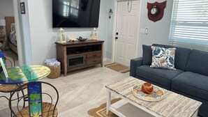 Un soggiorno con una TV a parete, un divano con un cuscino decorativo, un tavolino con un vassoio e una poltrona in vimini.