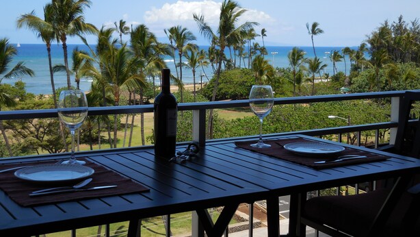 A table set for two with a bottle of wine and two glasses, overlooking a beach with palm trees and the ocean. - #605 Top Floor Unobstructed Ocean View/Sunsets,  New quiet A.C. (Kihei)