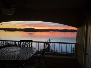 A balcony with a table and chairs overlooking a calm lake during sunset.