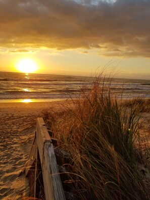 Une promenade en bois menant à une plage où l’on peut voir des vagues et un coucher de soleil en arrière-plan.