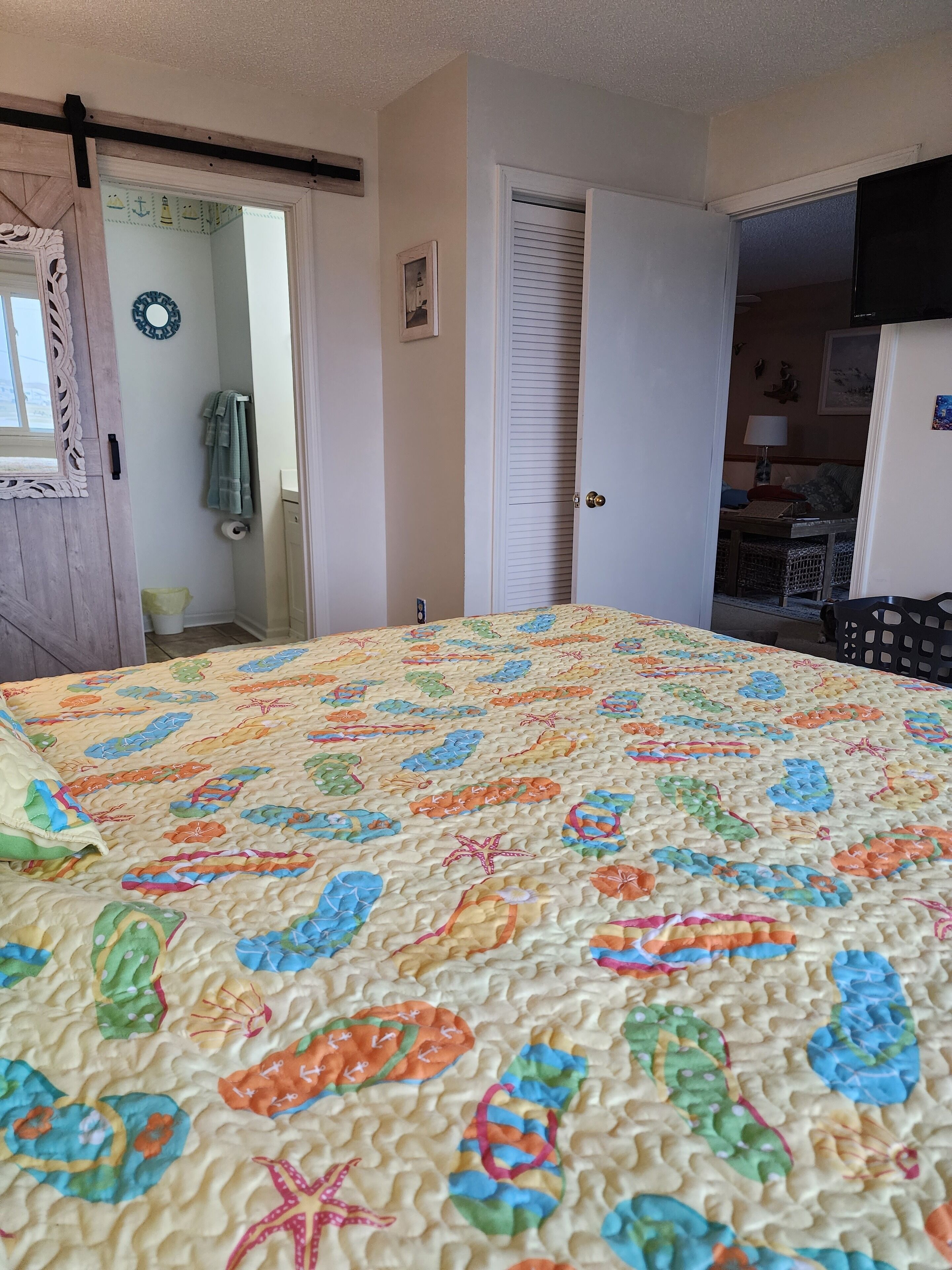 A bedroom with a bed covered in a colorful quilt, a wooden door, a white closet, and a view into another room with a table and chairs.