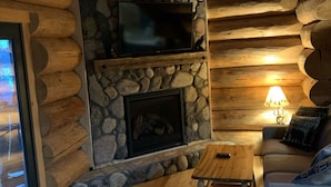 A cozy log cabin interior with a stone fireplace, a flat-screen TV mounted above it, a wooden coffee table, and a sofa with a cushion. - Log Cabin on beautiful Howard Lake. (Akeley)