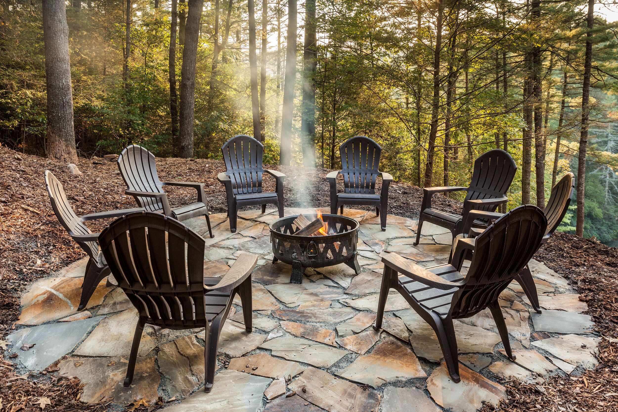 A stone fire pit surrounded by Adirondack chairs in a forest setting.
