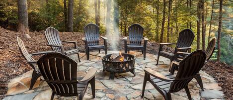 A stone fire pit surrounded by Adirondack chairs in a forest setting.