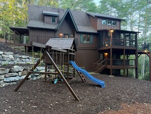 A two-story wooden house with a slide and a swing set in the backyard. - Gather & Unwind-Mountain Views, Hot Tub, Playground, River Fishing. Room for All (Ellijay)