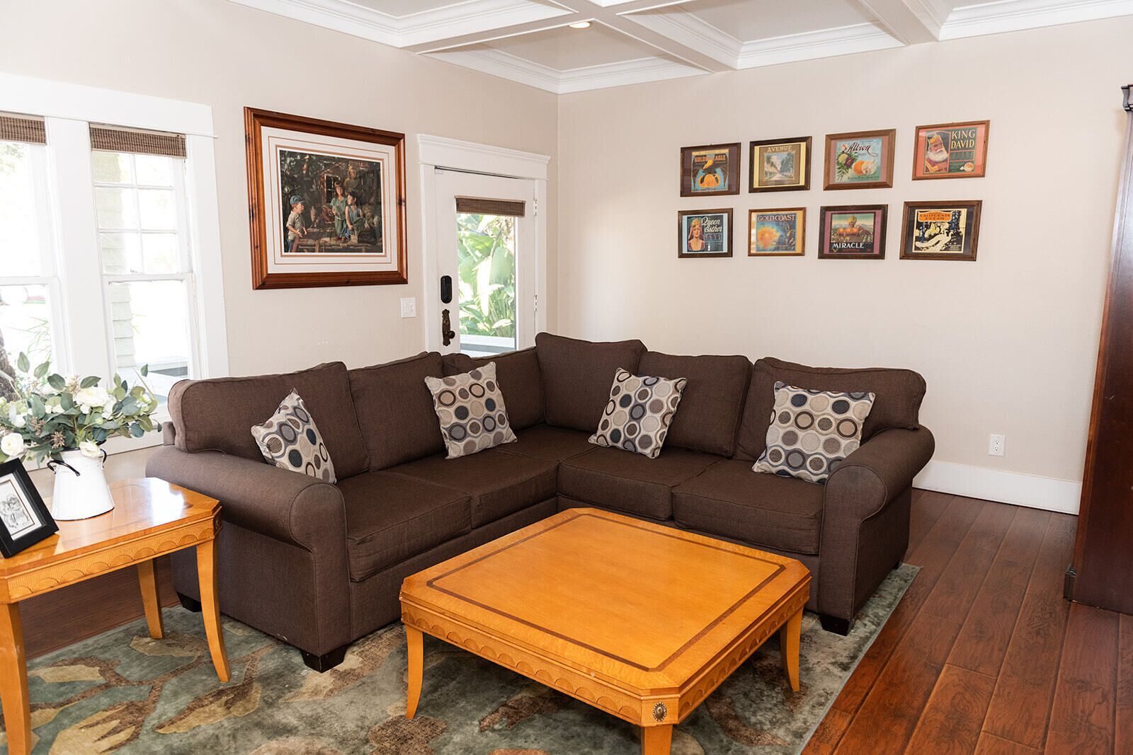 A living room with a brown sectional sofa, a wooden coffee table, and a rug. There are framed pictures on the wall and a vase with flowers on a side table.