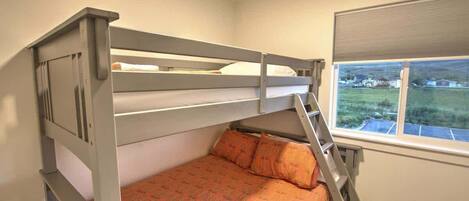Een stapelbed met een ladder, een bed met een decoratieve spreidek en een raam met uitzicht op een groen landschap.
