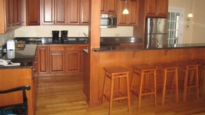 Eine Küche mit Holzschränken, einem Edelstahl-Kühlschrank, einer Mikrowelle und einer Kaffeemaschine. Am Tresen stehen drei Holzstühle.
