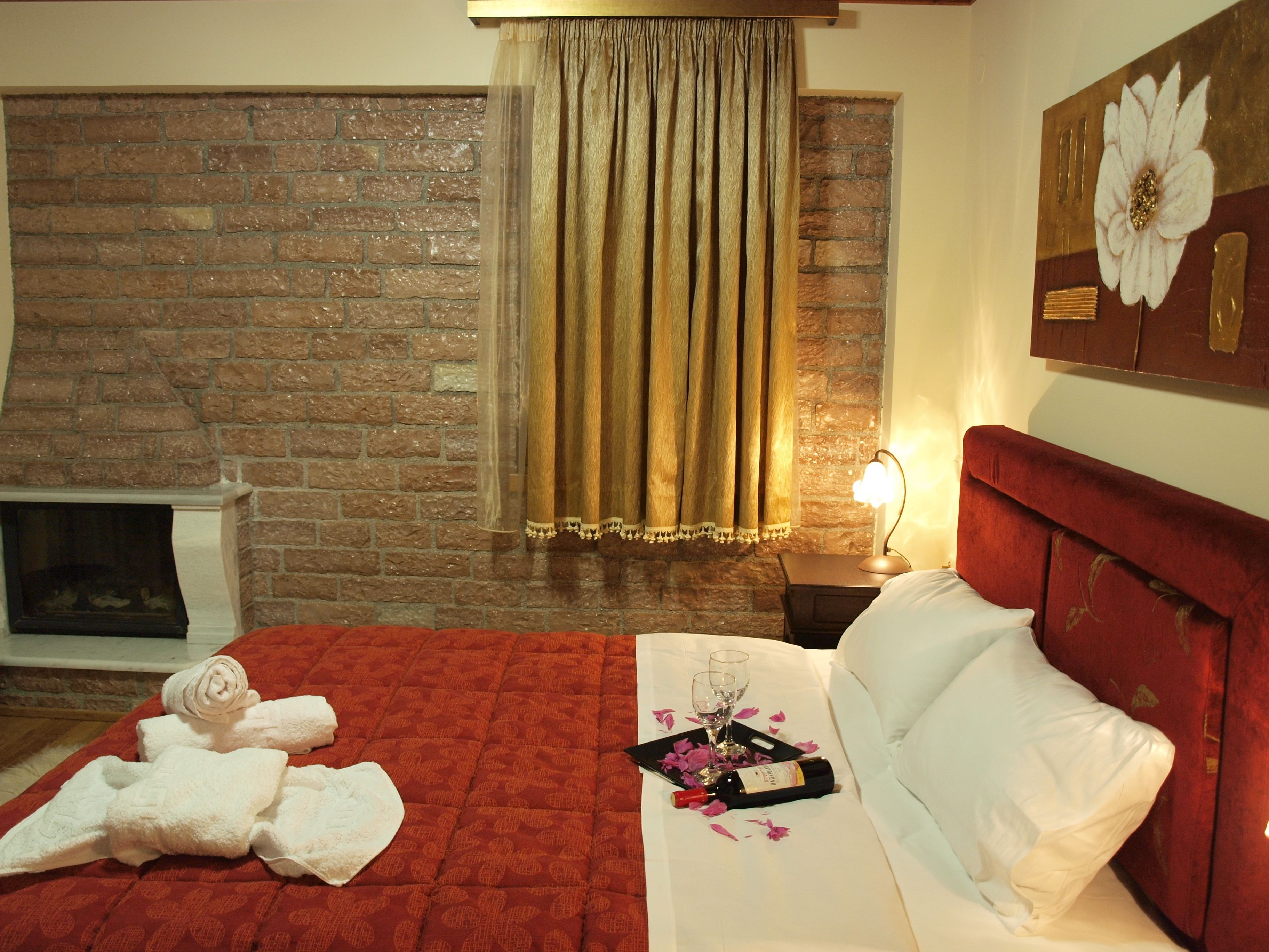 A hotel room with a brick fireplace, a bed with red headboard, a nightstand with a lamp, and a painting on the wall.