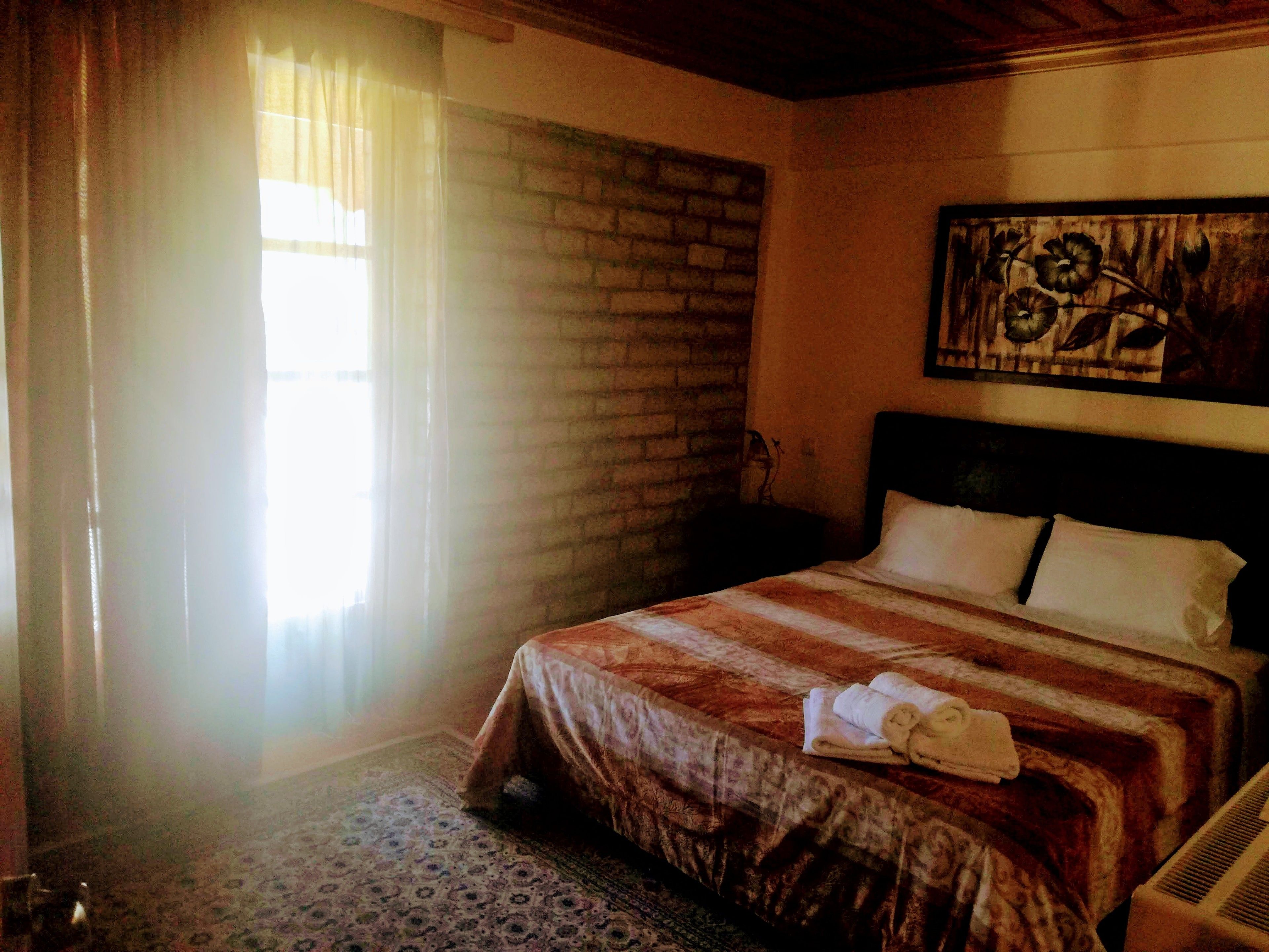 A hotel room with a bed, a nightstand, a painting on the wall, and a window with curtains.