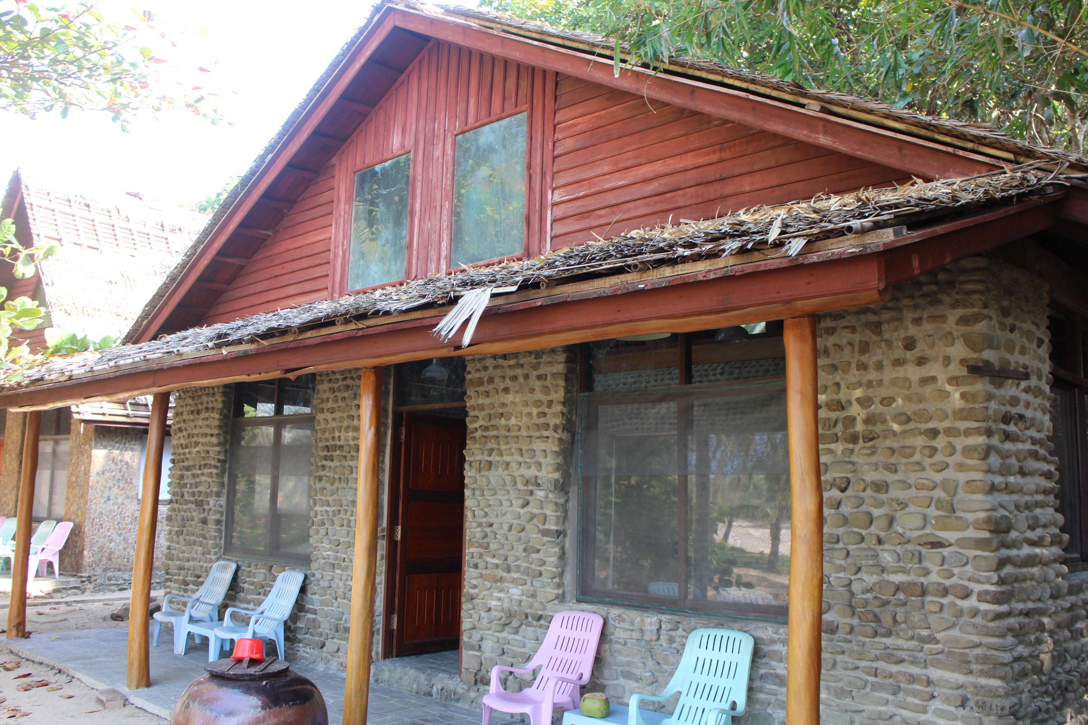 A rustic stone building with a red roof and a porch with plastic chairs.