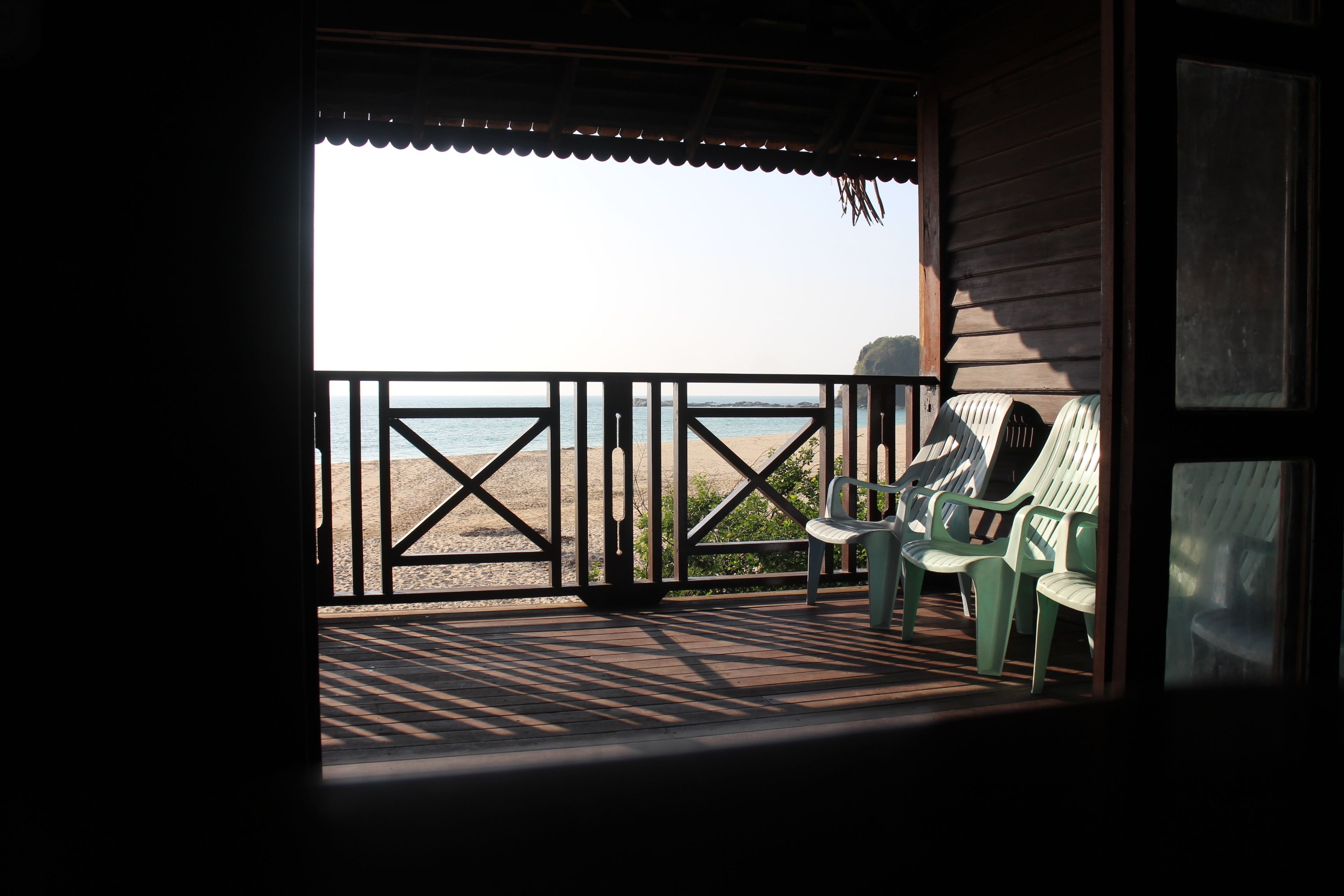 A wooden deck with a railing overlooking a beach. There are two green plastic chairs and a table on the deck.