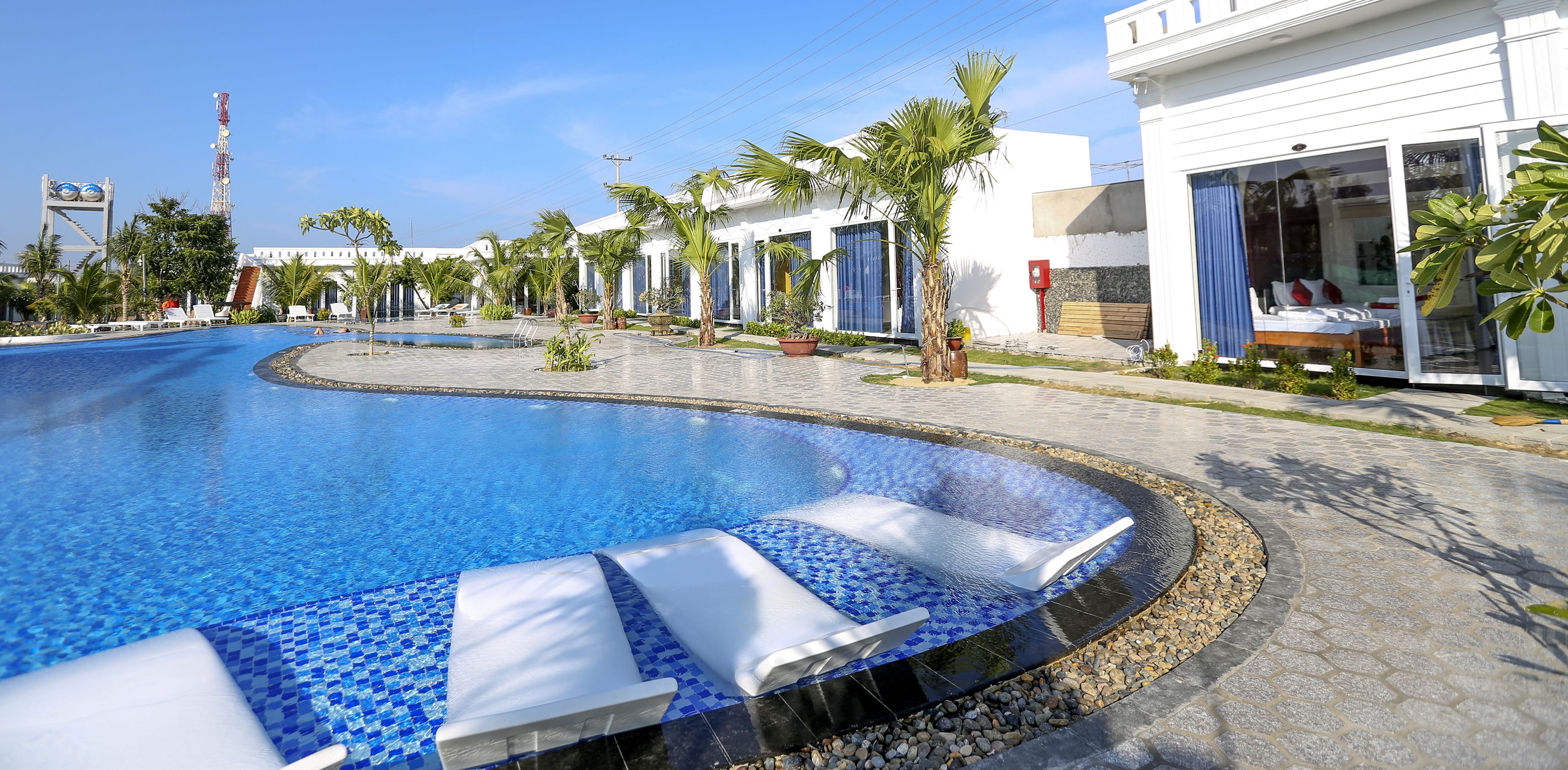 A swimming pool with lounge chairs, palm trees, and a white building with a blue door.