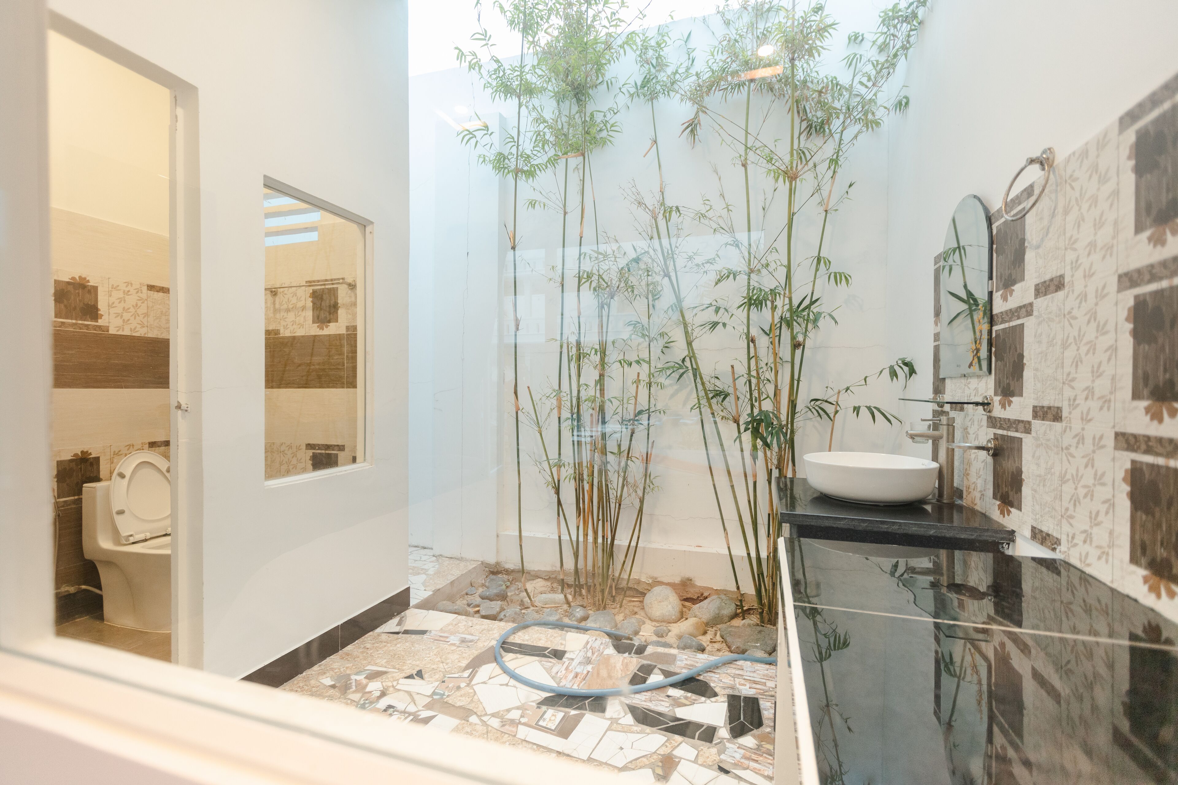 A bathroom with a toilet, a sink, and a mirror. There are bamboo plants and a hosed floor.