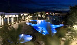 A nighttime view of a swimming pool area with a water feature, lounge chairs, and a building with lights.
