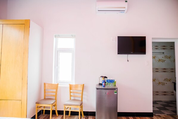 A room with a wooden wardrobe, a wooden chair, a small refrigerator with beverages, a television, and a window.