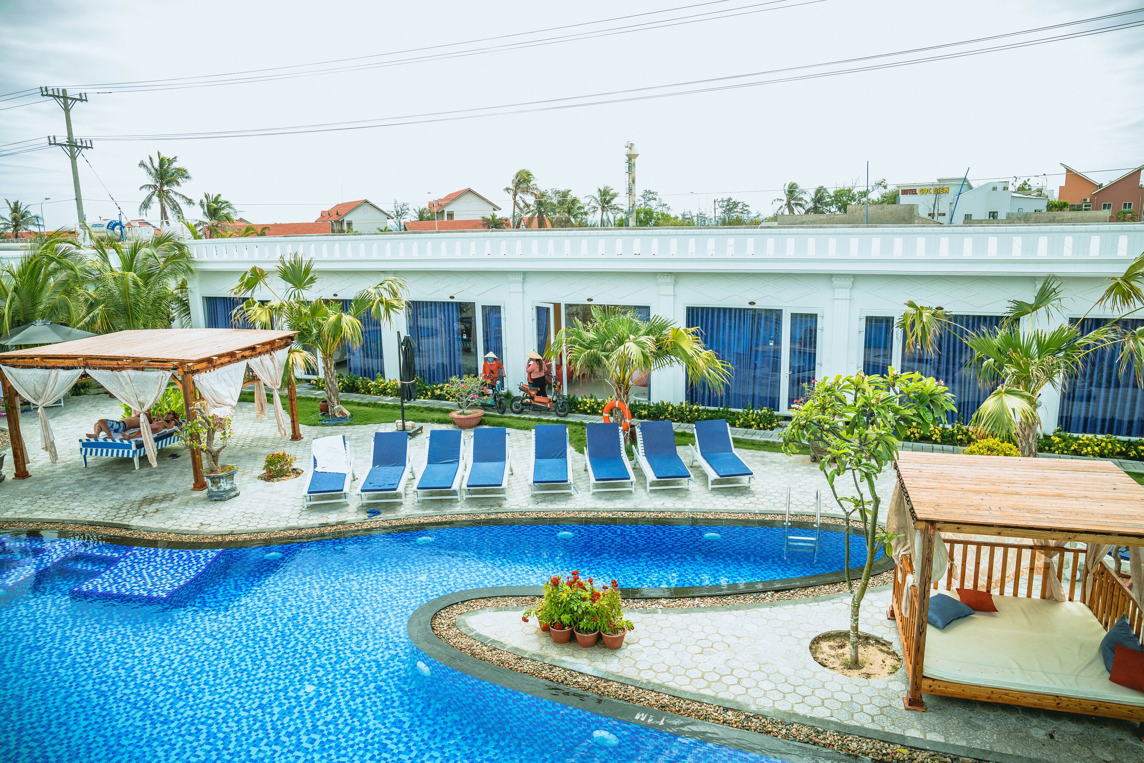 A swimming pool with lounge chairs, a wooden deck, and a building with blue doors in the background.