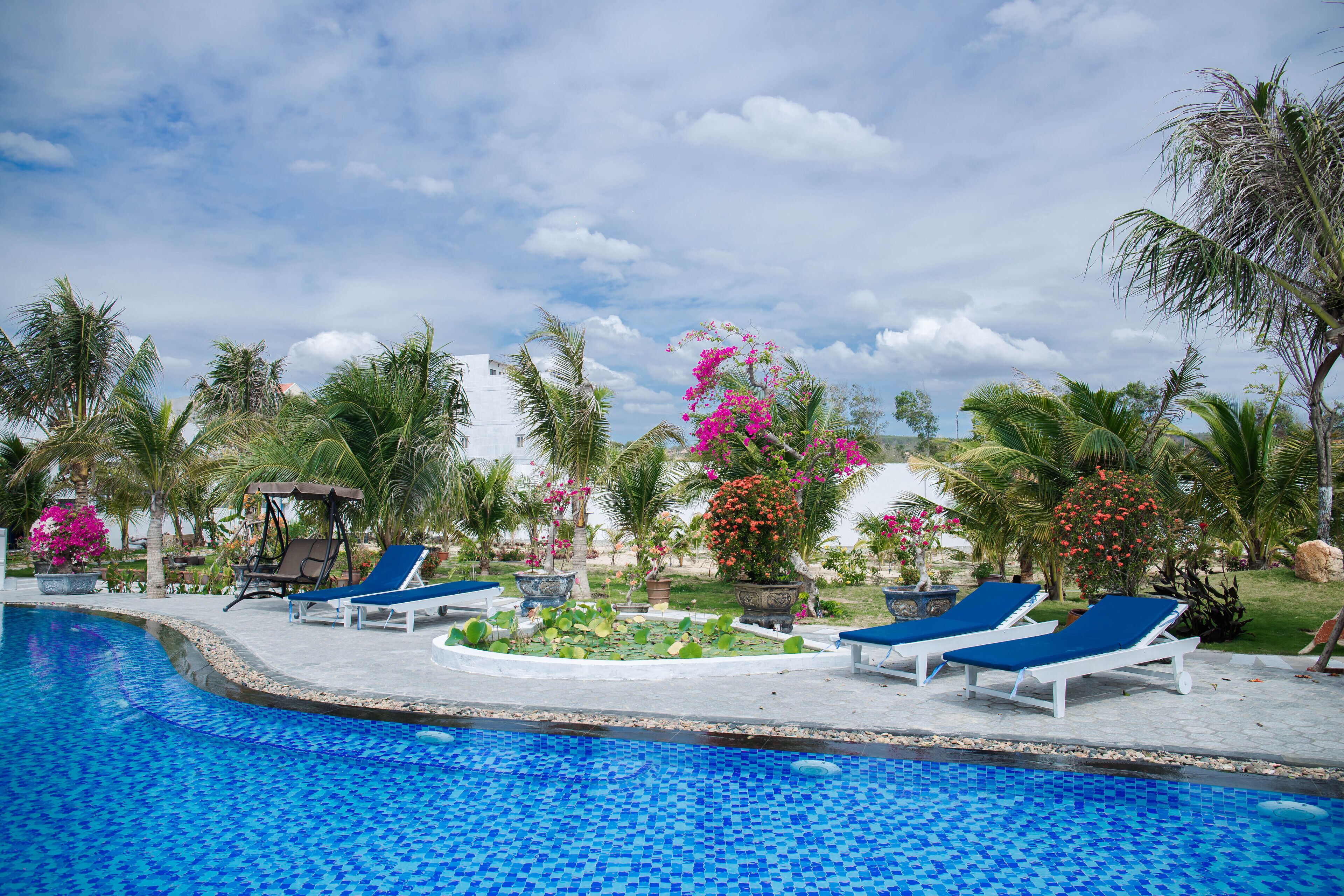 A swimming pool with blue mosaic tiles, surrounded by palm trees and various flowering plants.