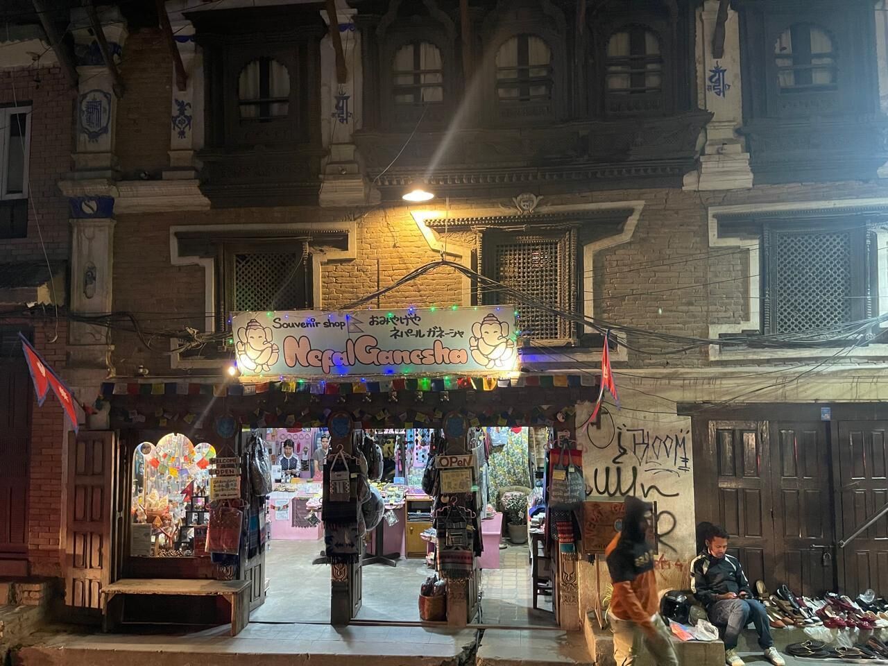 A traditional Nepalese shop with a signboard, various goods displayed outside, and a person sitting on a bench nearby.