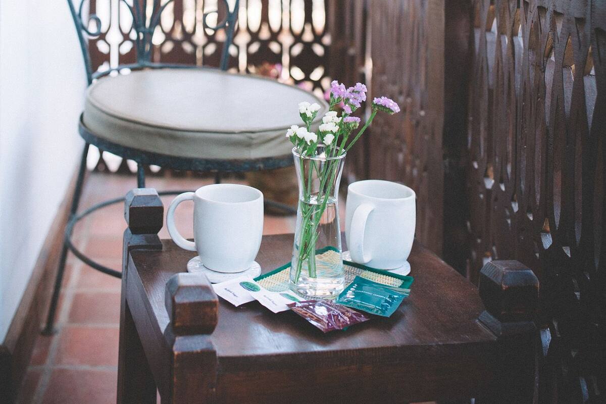A wooden table with two white cups and a vase of flowers on a balcony with a metal chair and a wooden railing.