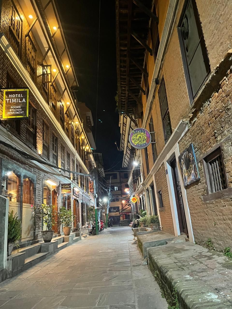 A narrow street lined with buildings, including a hotel named 'Hotel Timila' with rooms, studios, and apartments.