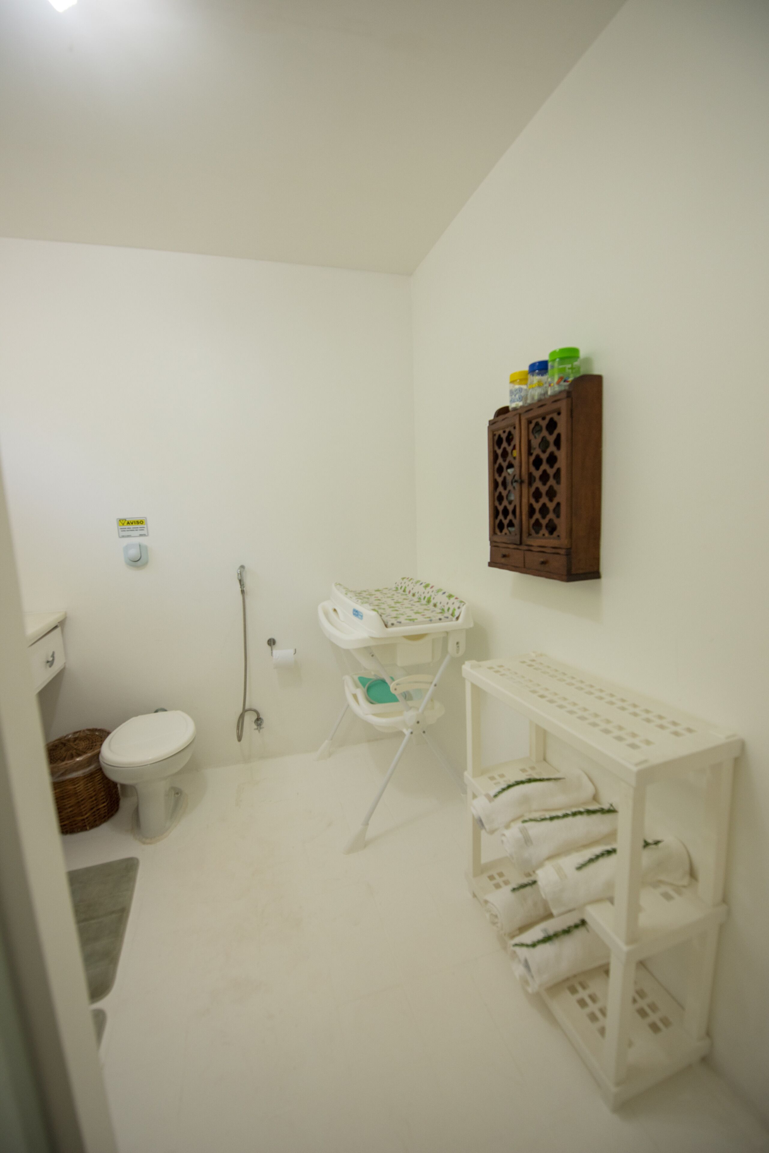 A small, white bathroom with a toilet, a sink, a cabinet with toiletries, and a shelf with folded towels.