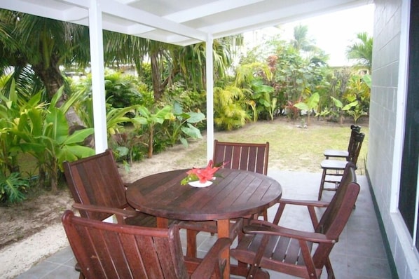 An outdoor patio with a wooden table and chairs, surrounded by lush tropical plants. - Lyas Holiday Home - 20 metres from a stunning beach (Tikioki Beach)