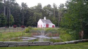 Ländliche Szenerie mit einem weißen Scheunenhaus, einem Teich mit Springbrunnen und einer Steinmauer.