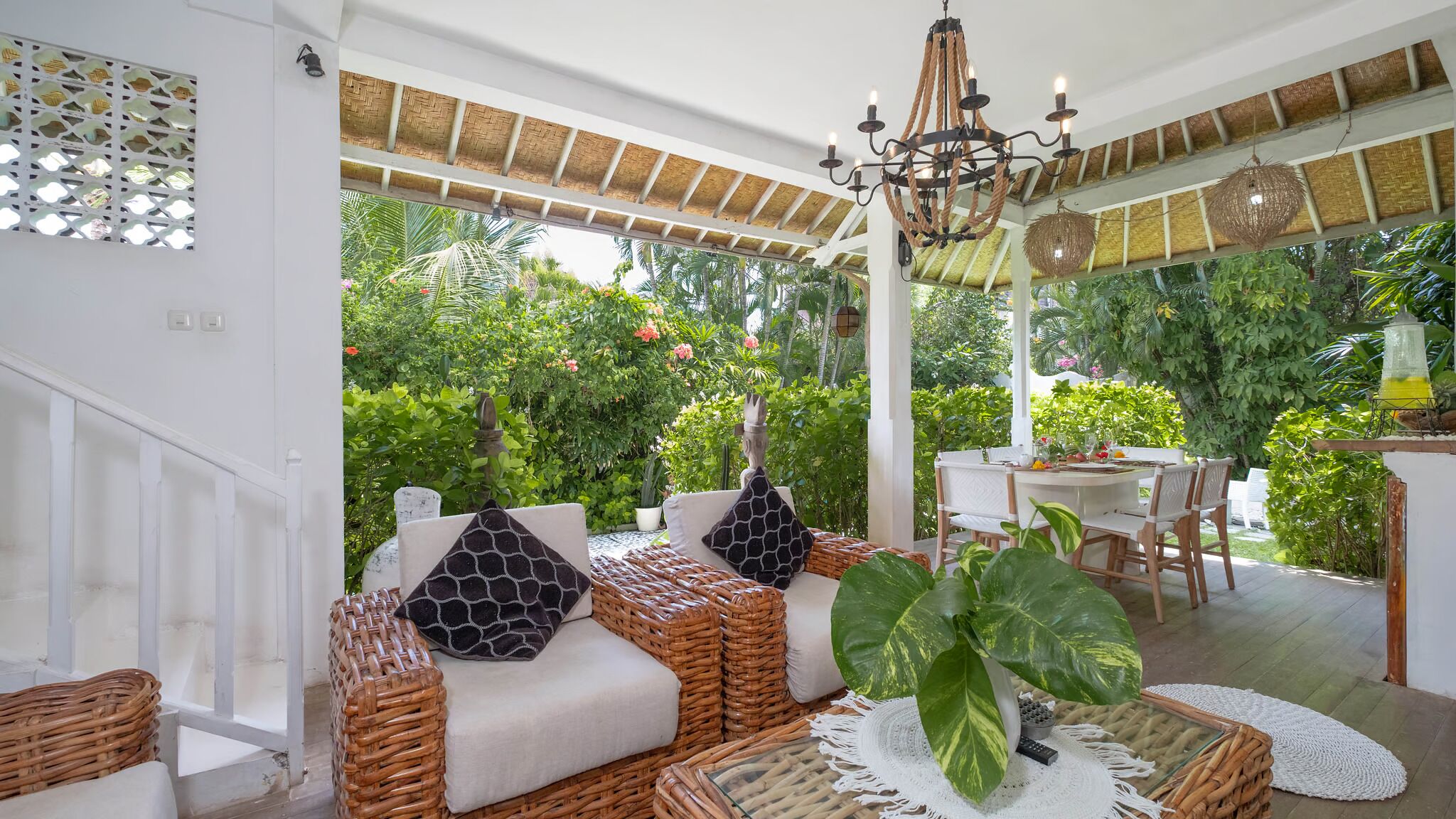 A covered outdoor seating area with wicker furniture, a dining table, and a large plant.