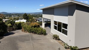 A modern two-story house with a garage, surrounded by greenery and a clear view of the cityscape and mountains in the distance. - Luxury Rotorua Executive Home with Lake Views: Relax, Unwind, and Enjoy (Rotorua Township)