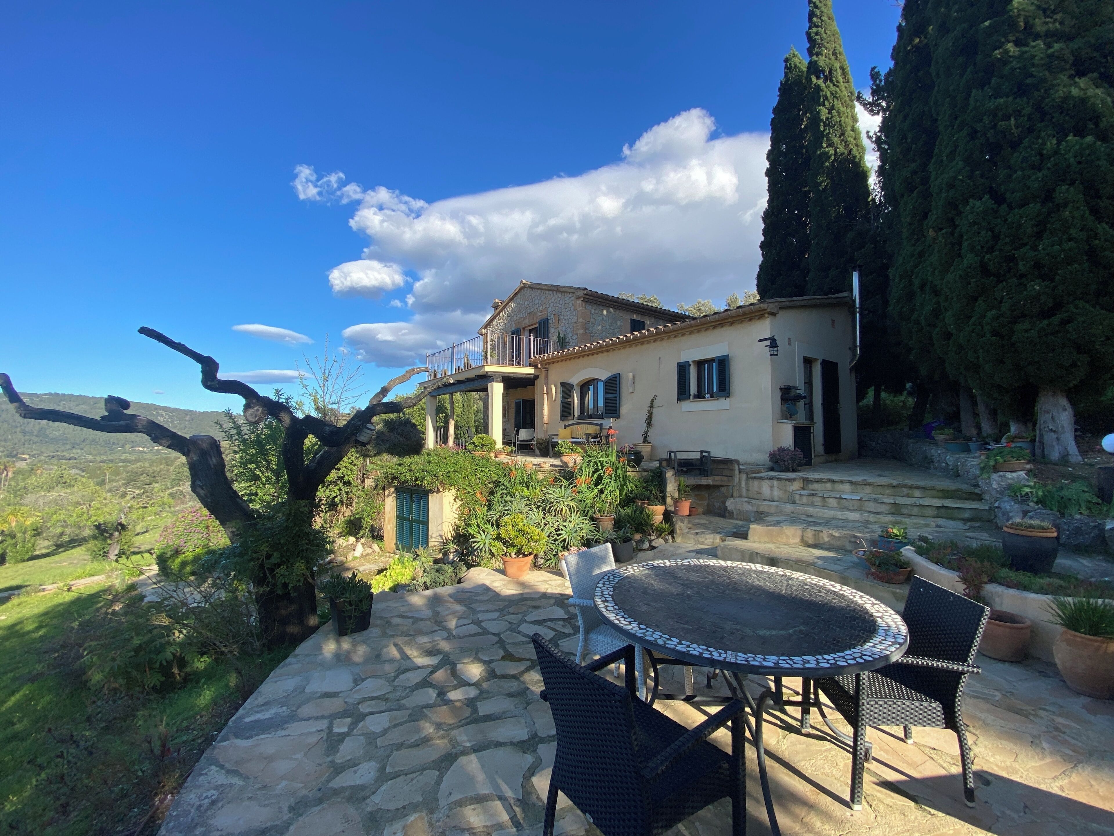 Un patio de piedra con una mesa redonda y sillas, una casa con balcón y vistas del paisaje circundante.