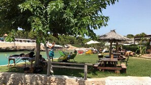 A park with picnic tables, a thatched-roof structure, and a body of water in the background.
