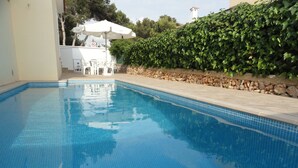A swimming pool with clear blue water, surrounded by a stone wall and a hedge. There is a white outdoor seating area with umbrellas in the background.