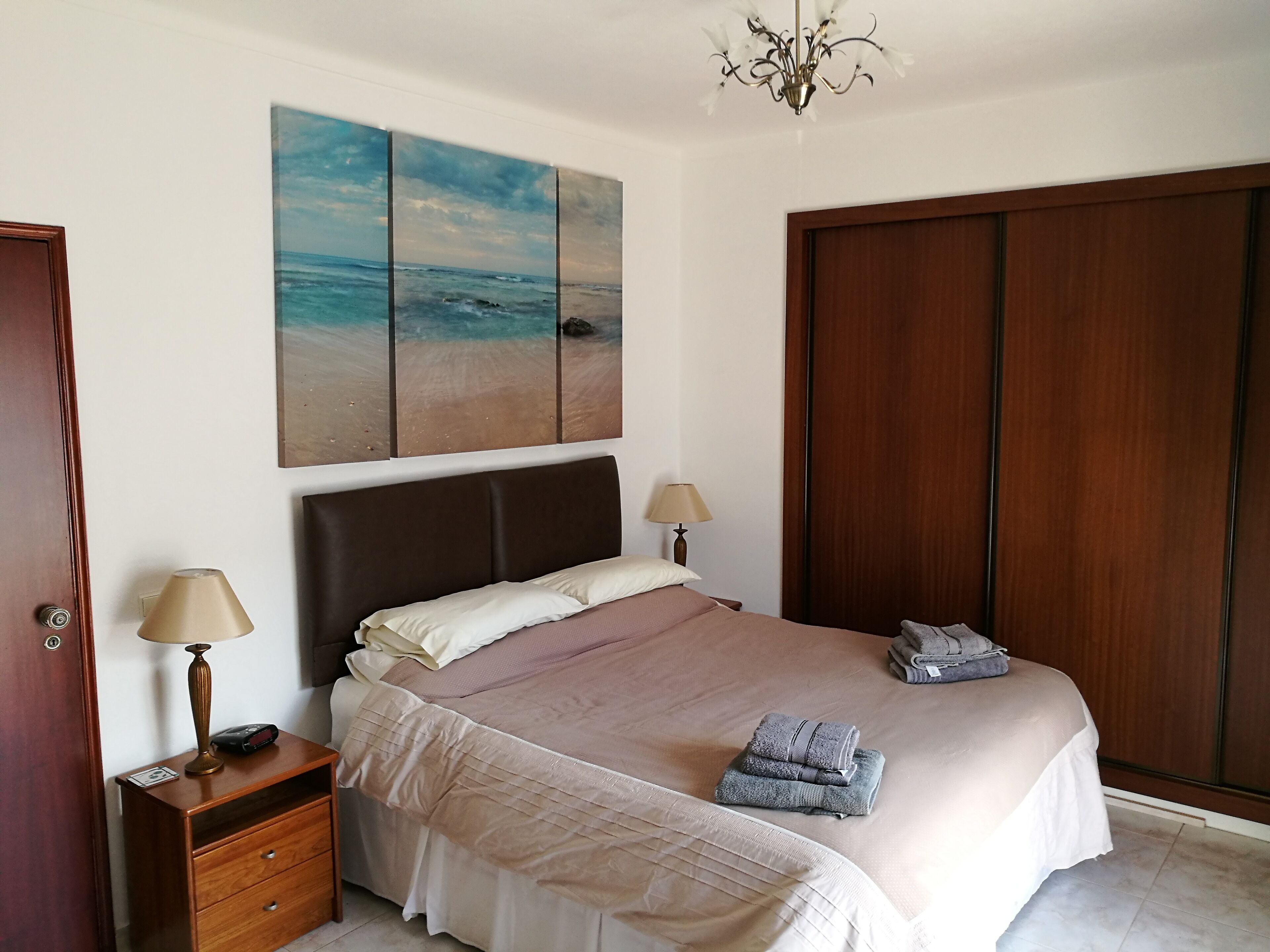Une chambre à coucher avec un lit, deux lampes de chevet, une table de chevet, une armoire en bois et un tableau sur le thème de la plage accroché au mur.