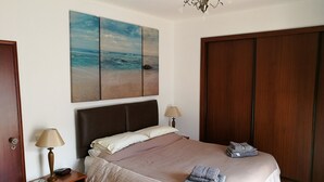 Een slaapkamer met een bed, twee bedlampen, een nachtkastje, een houten garderobe en een schilderij met een strandthema aan de muur.