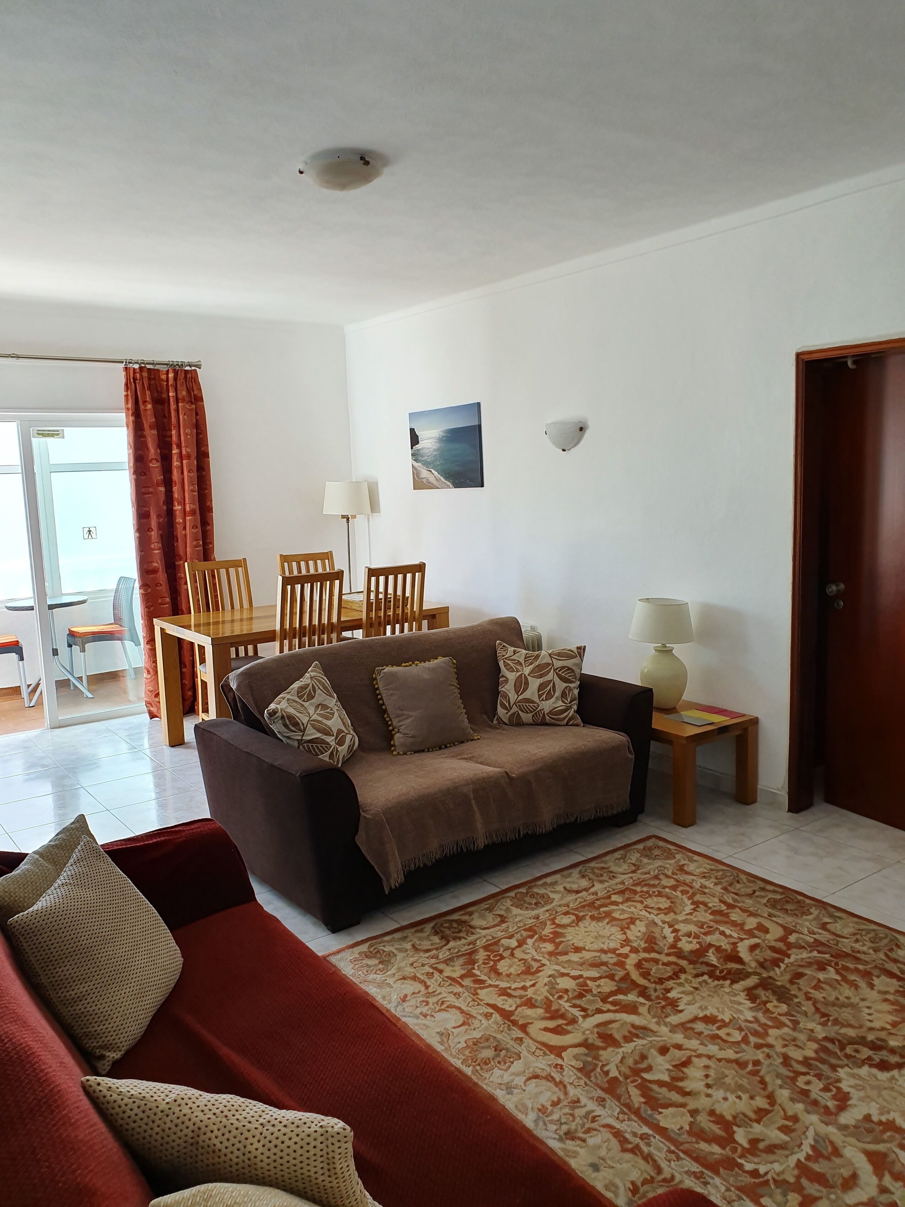 A living room with a brown sofa, red and beige patterned cushions, a wooden coffee table, and a patterned rug. There is a dining area with a wooden table and chairs, and a painting of a beach scene on the wall.