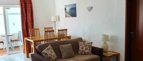 A living room with a brown sofa, red and beige patterned cushions, a wooden coffee table, and a patterned rug. There is a dining area with a wooden table and chairs, and a painting of a beach scene on the wall.