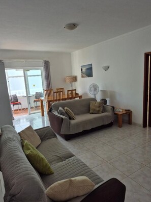 A living room with a sofa, a dining table with chairs, a fan, and a view through a glass door.