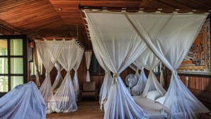 Ein Zimmer mit vier Betten, die mit weißen Baldachinen versehen sind, Holzböden und einem Ventilator in der Mitte.