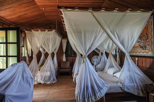 Habitación con cuatro camas cubiertas por doseles blancos, piso de madera y un ventilador en el centro.