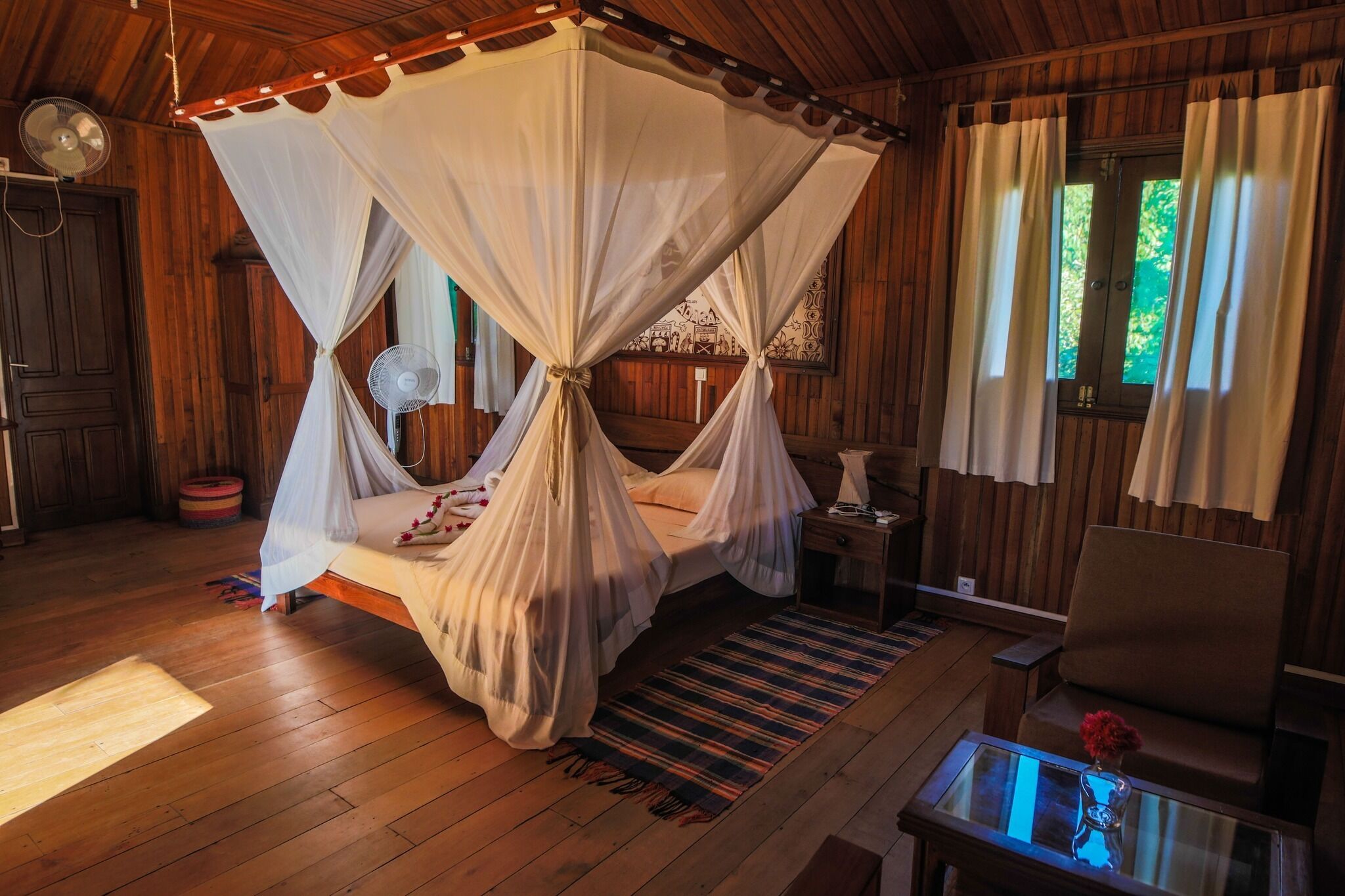 A wooden room with a four-poster bed, a fan, and a small table with a vase.