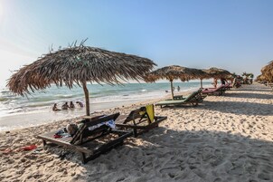 Una playa con sombrillas de paja, sillas de descanso y gente en el agua.