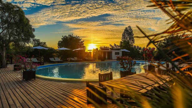 Ein Poolbereich mit Liegestühlen und Sonnenschirmen, umgeben von Bäumen und Blick auf einen Sonnenuntergang.