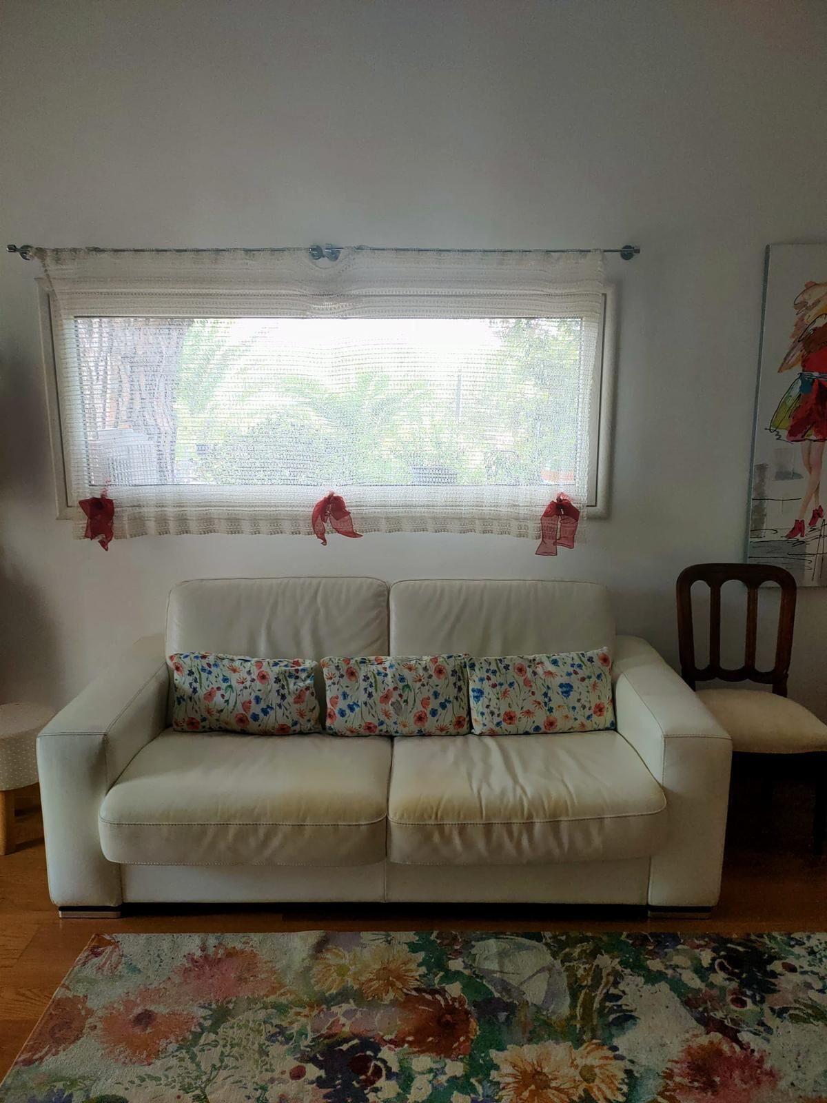 Un salon avec un canapé blanc, des coussins à motifs floraux, un tapis à dessins, une chaise en bois et une fenêtre ornée de rideaux légers.