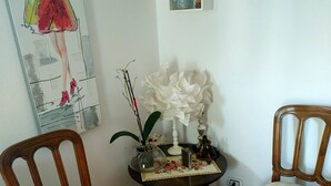 A room with a wooden chair, a small table with decorative items, a wall-mounted shelf with figurines, and a painting.