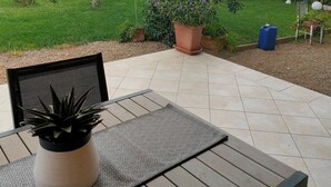 A patio with a table and chairs, surrounded by potted plants and a view of a lush garden and a building in the background.