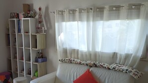 A white leather sofa with a red pillow, a white bookshelf, and a window with sheer curtains.