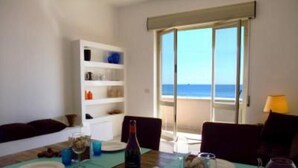 A dining area with a table set for two, a bookshelf, and a view of the sea through a large window.