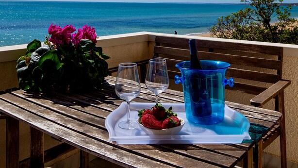 A wooden table with a white cloth, two wine glasses, a blue water container, and a plate with strawberries on a balcony overlooking the sea.