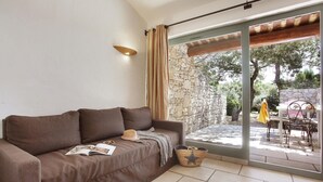A brown sofa with a magazine, a straw hat, and a woven basket in a room with a stone wall and a glass door leading to an outdoor area with a table and chairs. - Maisonette in Gordes with Provençal Charm (GORDES)